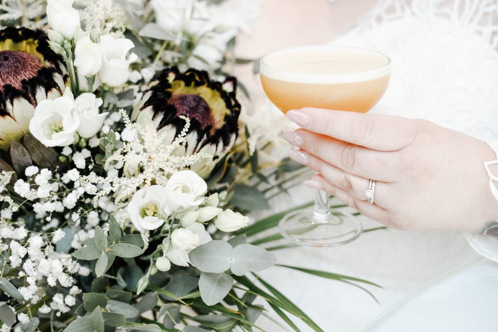 Bride's hand holding the glass with wedding drink showing her wedding ring and flowers bouquet